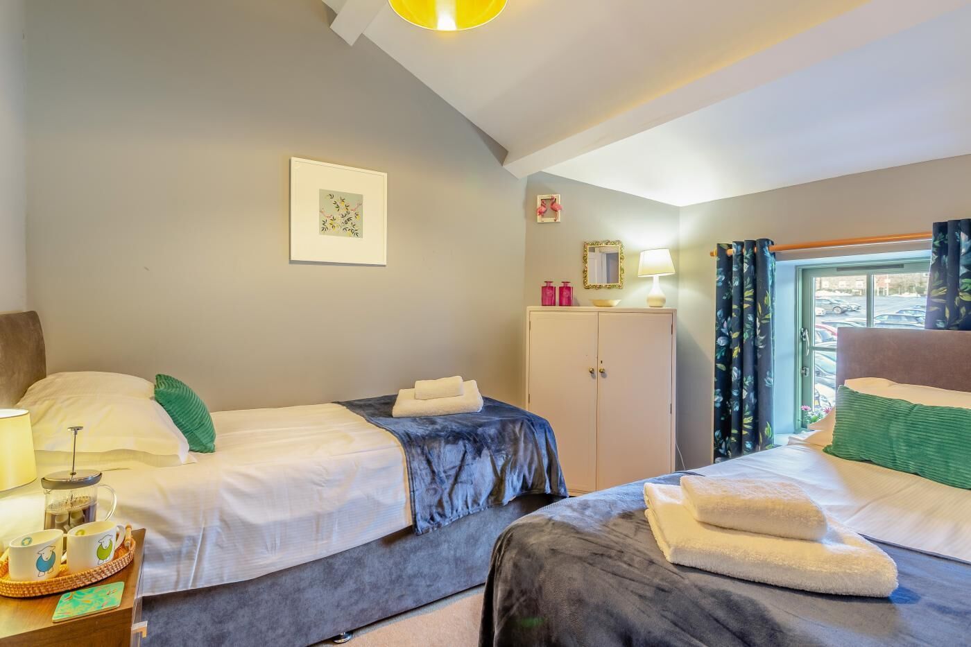 Een hotelkamer met twee bedden, een kledingkast, een nachtkastje met een lamp, een raam met gordijnen en een ingelijste foto aan de muur.