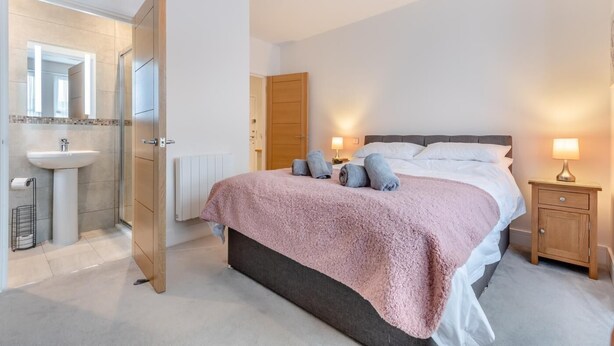 A bedroom with a bed, bedside lamps, a nightstand, a wooden door, a bathroom with a sink and mirror, and a shower area. - 2 Bed in Swanage (oc-w30658) (Swanage)