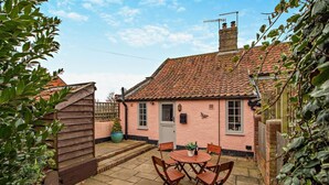 Une maison rose avec un toit en tuiles, un abri en bois et une terrasse avec une table et des chaises.