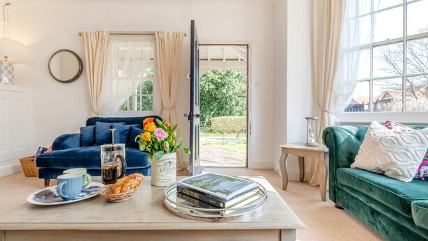 Ett vardagsrum med en blå soffa, en grön soffa, ett soffbord med böcker och en vas med blommor, och en dörr som leder ut till en trädgård.