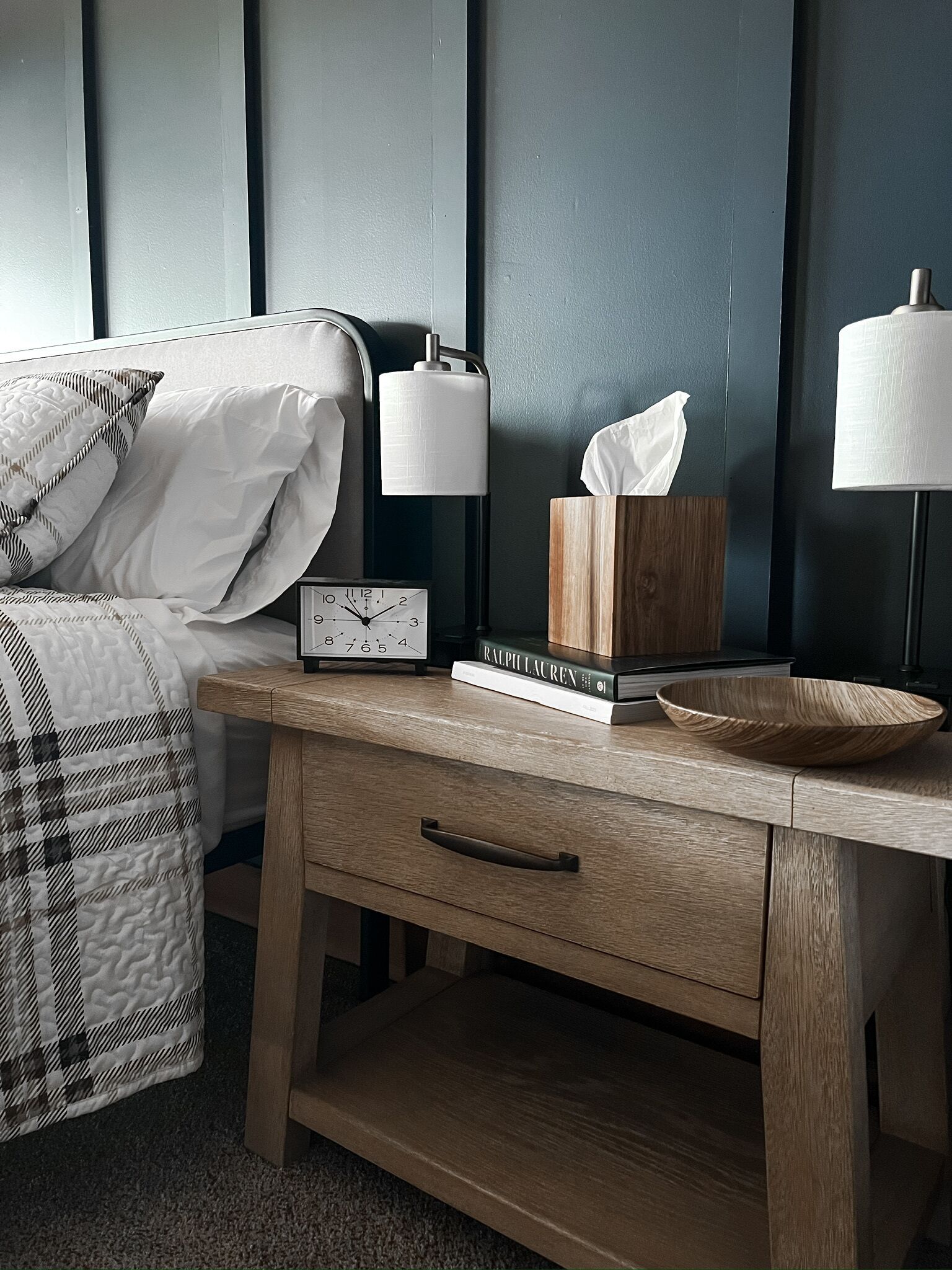 A neatly made bed with a patterned blanket, a bedside table with a clock, a tissue box, and a bowl.