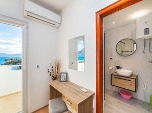 Ein modernes Badezimmer mit einem großen Spiegel, einem Waschbecken und einer Toilette. Ein Holztisch mit Bilderrahmen und einer Vase mit Trockenpflanzen. Das Badezimmer bietet Blick auf das Meer und die Berge.