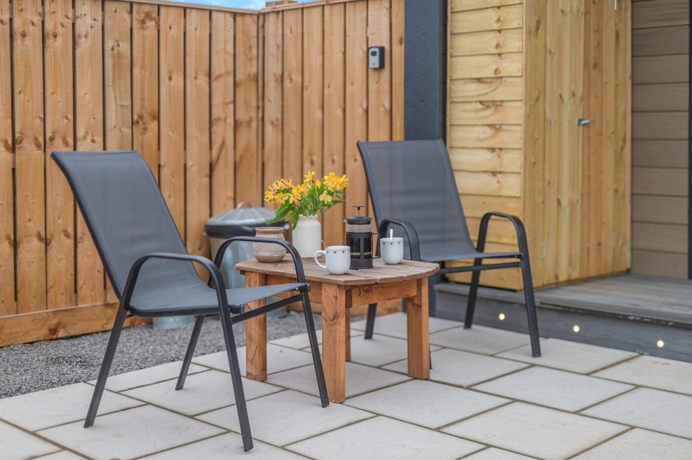 Een houten terras met twee zwarte stoelen en een kleine tafel, een vaas met bloemen en een koffiezetapparaat.