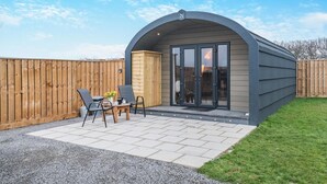 Un pequeño patio exterior moderno con una estructura de techo curva, una disposición de asientos con dos sillas y una mesa, y una valla de madera al fondo.