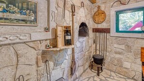 Una habitación rústica con paredes de piedra, techo de madera, un cuadro enmarco de 'La Última Cena', un botellero de madera, un hogar de piedra y una ventana con vistas a follaje.