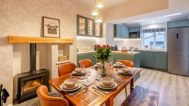Comedor con mesa de madera preparada para cuatro personas, chimenea y cocina al fondo.