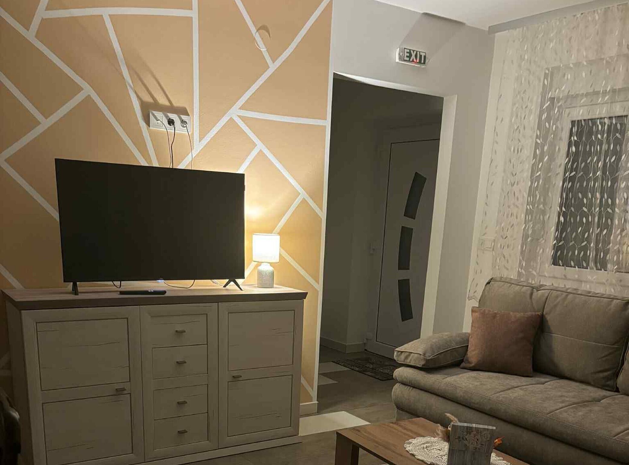Una sala de estar con un televisor de pantalla plana sobre un mueble, un sofá, una lámpara y una señal de salida.