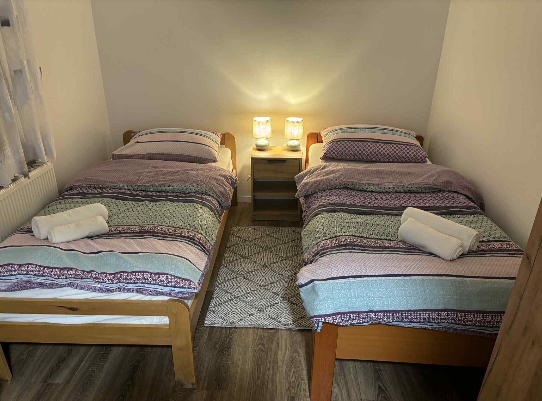 Una habitación pequeña y acogedora con dos camas individuales, una mesita de noche con dos lámparas y una alfombra estampada.