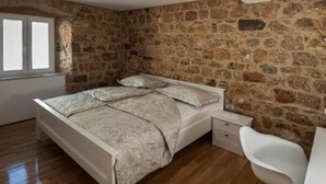 Une chambre avec un mur en pierre, un plancher en bois, un lit avec une tête de lit blanche, une table de chevet et une chaise blanche.