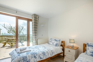 A bedroom with a bed, bedside tables, a lamp, a view of the outdoors, and a window with curtains. - 4 Bed in St. Endellion (oc-t30725) (Trelights)