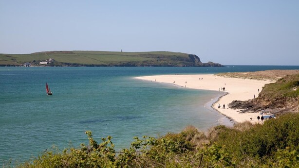 Une plage côtière offrant une vue dégagée sur la mer, une étendue de sable et, au loin, une colline surmontée d’un phare.