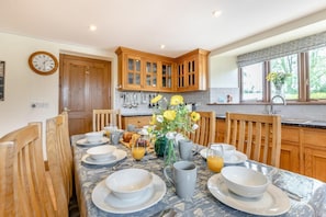 A dining area with a table set for breakfast, wooden cabinets, a clock, and a window with a view. - 4 Bed in St. Endellion (oc-t30725) (Trelights)