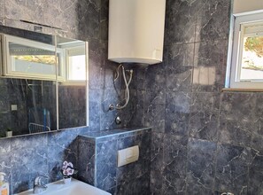 A bathroom with dark marble tiles, a large mirror, a wall-mounted light fixture, and a shower area. - Apartment Beleca - One Bedroom Apartment with Balcony (Podstrana)