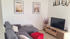 Un salon moderne avec un canapé d’angle gris, un pouf rouge, un meuble TV en bois et deux tableaux encadrés accrochés au mur.