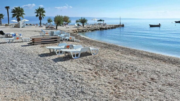 Une plage de galets avec des chaises longues blanches et des parasols, des palmiers et des bateaux sur l’eau.