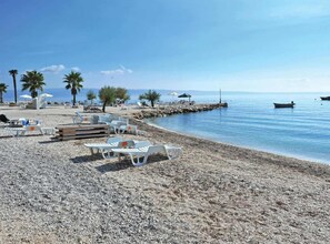 En strand med småsten, vita solstolar och parasoller, palmer och båtar i vattnet.
