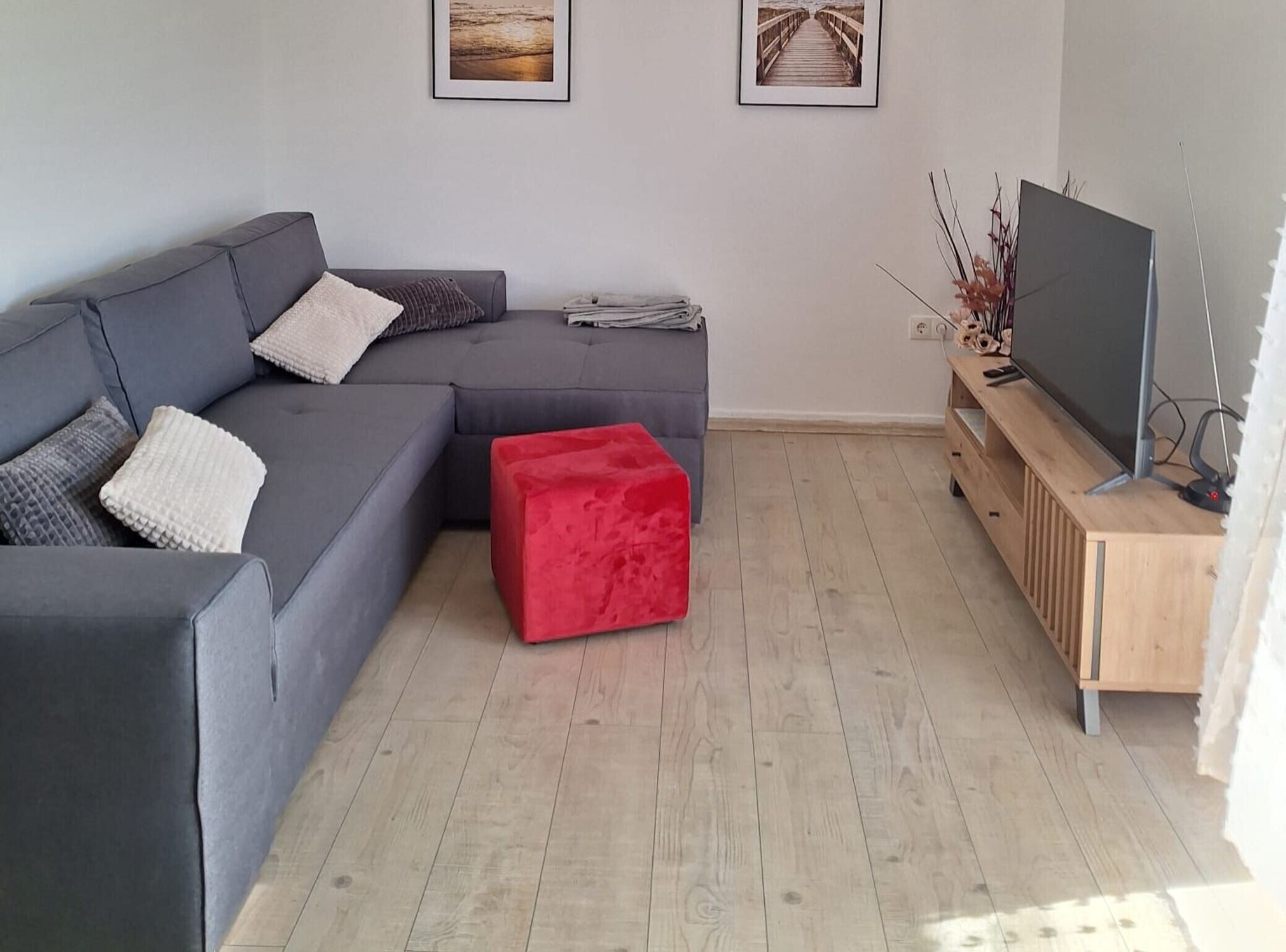 A modern living room with a grey sectional sofa, a red ottoman, a wooden TV stand, and two framed pictures on the wall.