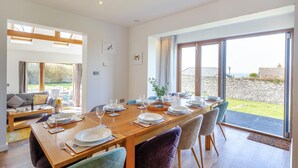 A dining area with a wooden table set for six, large windows, and a view of the sea. - 5 Bed in Trevone (oc-s30762) (Trevone)