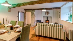 Un salon chaleureux avec une cheminée, un canapé à rayures, des poutres en bois et un coin repas avec une table mise pour le dîner.
