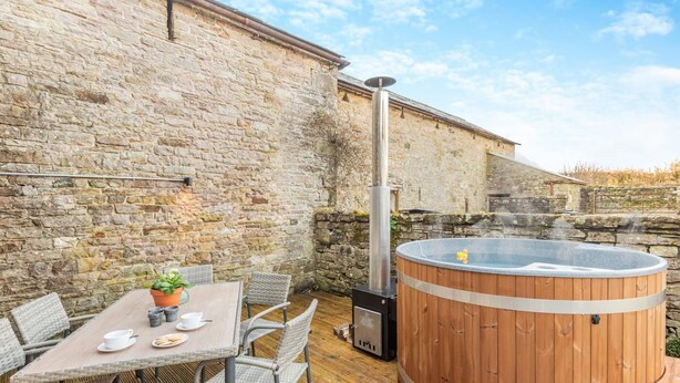 Un bâtiment en pierre avec un bain à remous en bois, une table avec des chaises et un pot de fleurs.