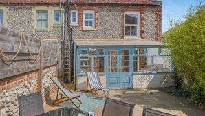 Une maison avec une façade en pierre, une extension dotée de fenêtres bleues et un espace extérieur aménagé avec des chaises et une table.