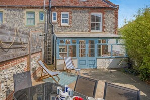 Une maison avec une façade en pierre, une extension dotée de fenêtres bleues et un espace extérieur aménagé avec des chaises et une table.