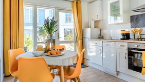Eine helle Küche mit weißen Schränken, ein Essbereich mit orangefarbenen Stühlen und ein mit Speisen gedeckter Tisch.