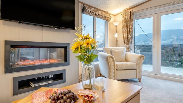A modern living room with a fireplace, a flat-screen TV, a comfortable armchair, and a large window offering a scenic view. - 2 Bed in St. Fillans (oc-l30650) (Saint Fillans)