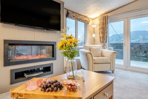 A modern living room with a fireplace, a flat-screen TV, a comfortable armchair, and a large window offering a scenic view. - 2 Bed in St. Fillans (oc-l30650) (Saint Fillans)