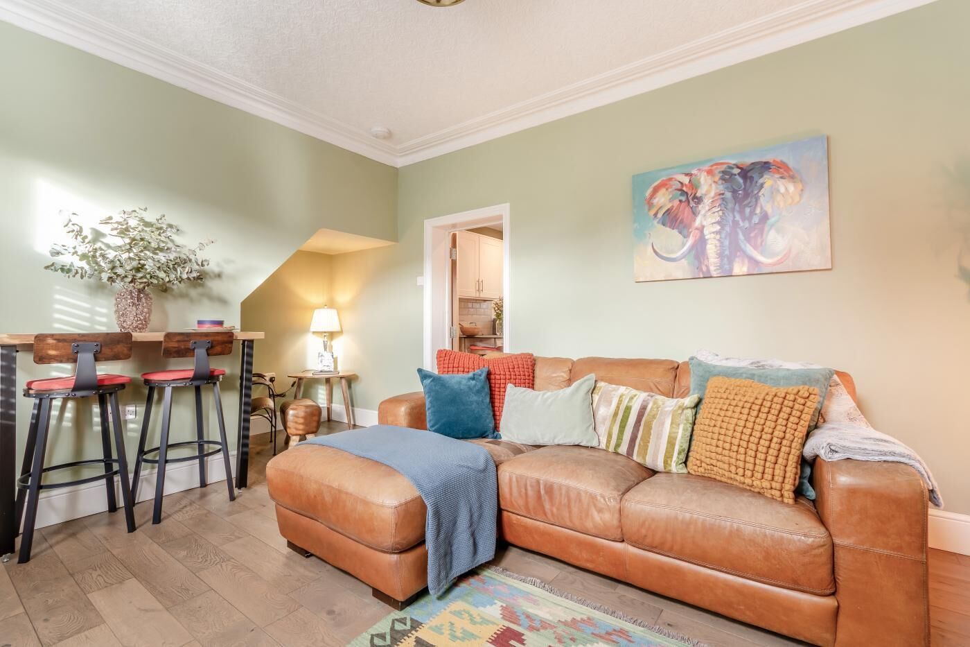 A living room with a brown leather sofa, two red bar stools, a lamp, a painting of an elephant, and a wooden floor.