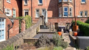 Ein traditionelles Backsteingebäude mit einer Steintreppe, die zu einer Haustür führt, einer kleinen Außenbereich-Sitzgelegenheit mit Stühlen und Tischen sowie einem Garten mit einer Steinsäule und Pflanzen.