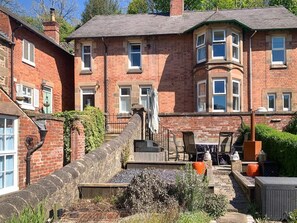 Ein traditionelles Backsteingebäude mit einer Steintreppe, die zu einer Haustür führt, einer kleinen Außenbereich-Sitzgelegenheit mit Stühlen und Tischen sowie einem Garten mit einer Steinsäule und Pflanzen.