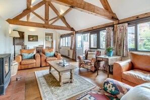 Un vaste salon doté de hauts plafonds, de poutres apparentes, de canapés en cuir, d’une table basse en bois et d’une grande fenêtre agrémentée de rideaux.