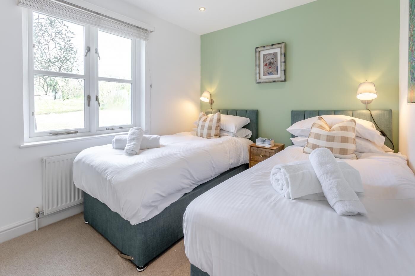 A hotel room with two beds, white bedding, a window with blinds, a radiator, and a framed picture on the wall.