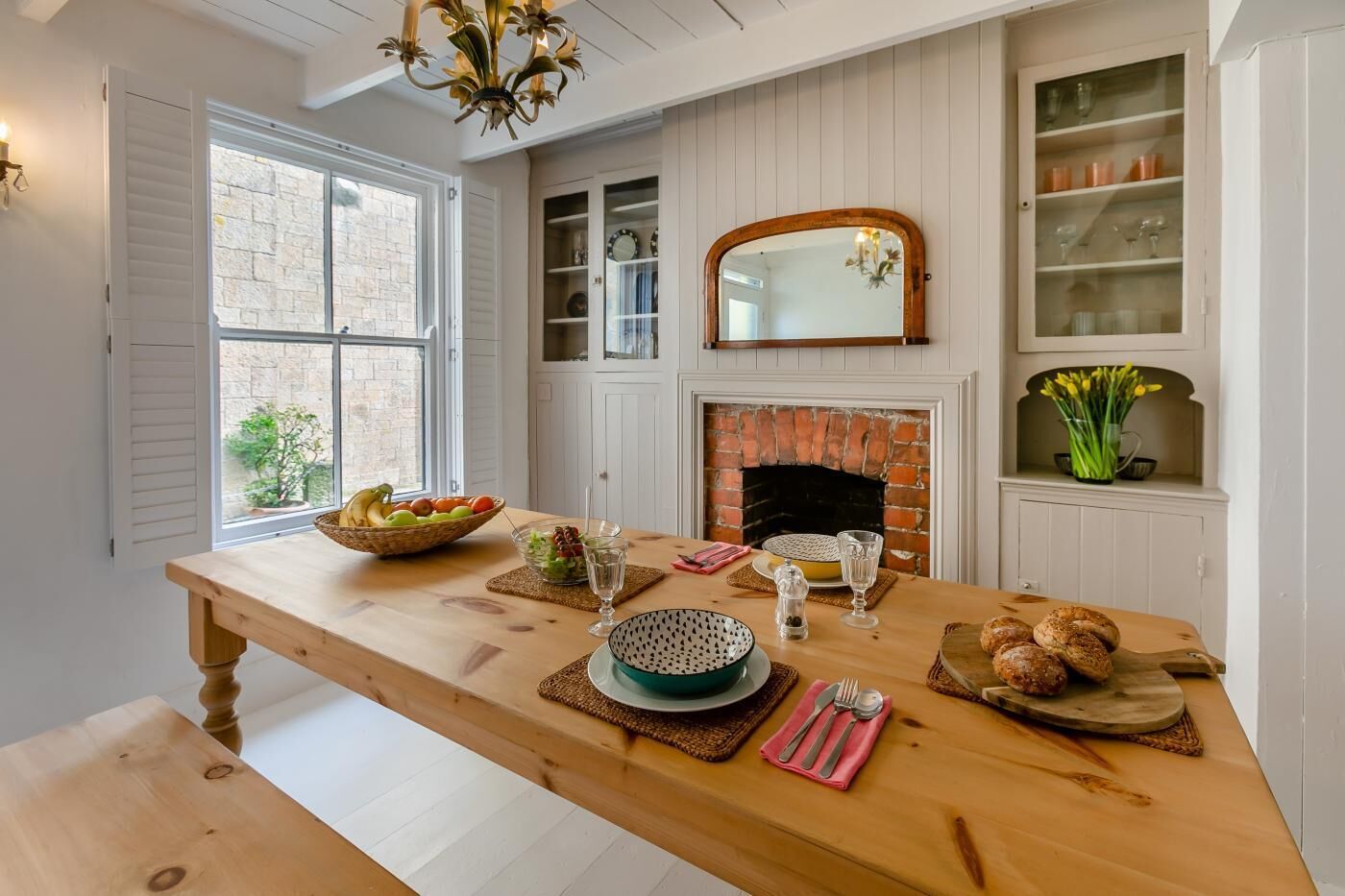 A dining area with a wooden table, a brick fireplace, and a window with shutters.
