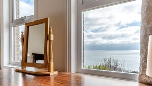 Een spiegel met houten lijst op een houten tafel, bij een groot raam met uitzicht op zee.