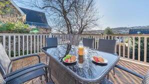 An outdoor dining area with a glass table, wicker chairs, and a view of a house and trees. - 3 Bed in Saundersfoot (oc-c30698) (Saundersfoot)