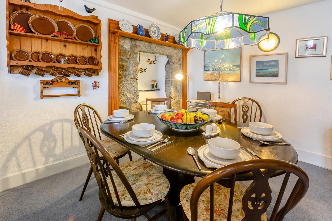 A dining area with a round table set for four, surrounded by chairs with floral upholstery. A large bowl of fruit is placed on the table, and there are plates and cutlery set.
