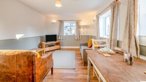 Un salon chaleureux avec un canapé en bois, un téléviseur posé sur un meuble en bois, une table à manger avec des couverts et une fenêtre avec des rideaux.