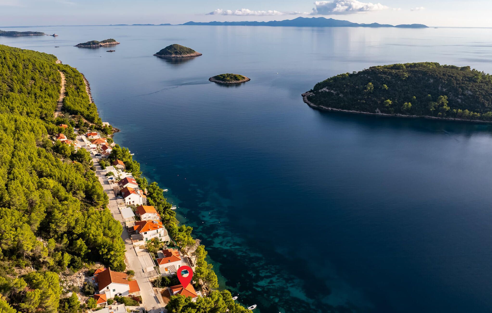 An aerial view of a coastal village with houses along the shoreline, clear blue sea, and small islands in the distance.