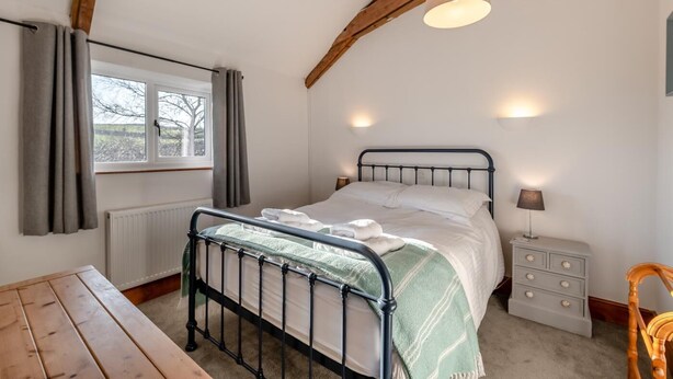 A bedroom with a metal bed frame, a bedside table, a lamp, and a window with curtains. - 3 Bed in East Allington (oc-m30710) (East Allington)