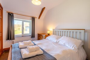 A bedroom with a large bed, a nightstand with a lamp, a window with curtains, and a radiator. - 4 Bed in East Allington (oc-k30713) (East Allington)