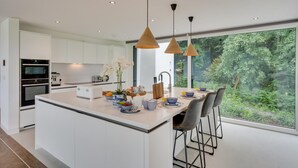 Een moderne keuken met witte kasten, een centraal eiland, hangende lampen en een groot raam met uitzicht op groen.