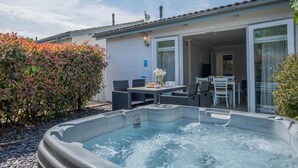Un jacuzzi situé devant une maison, avec une table et des chaises sur une terrasse, un espace extérieur couvert et une haie.