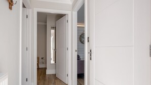 Un couloir avec des portes blanches menant à une pièce où se trouve un lit violet et une horloge au mur.