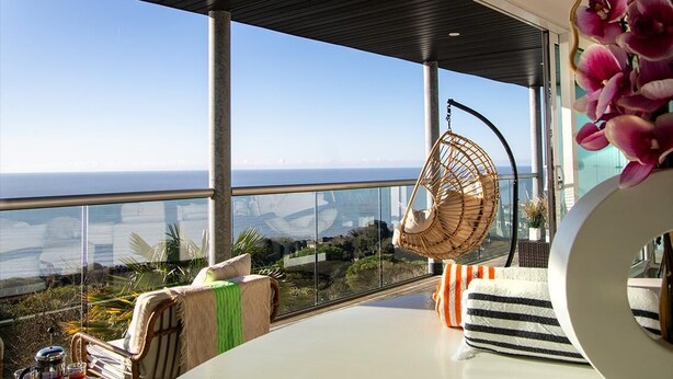 Una habitación moderna con un amplio ventanal que ofrece una vista panorámica, una silla de mimbre y una mesa blanca con cojines a rayas.