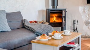 A cozy living room with a sofa, a table with pastries and cups, and a wood-burning stove. - 2 Bed in Clevedon (oc-s30785) (Clavedon)