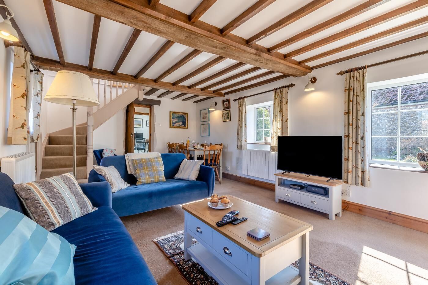 A cozy living room with a blue sofa, a wooden coffee table, and a television. A staircase leads to another room, and there is a window with curtains.