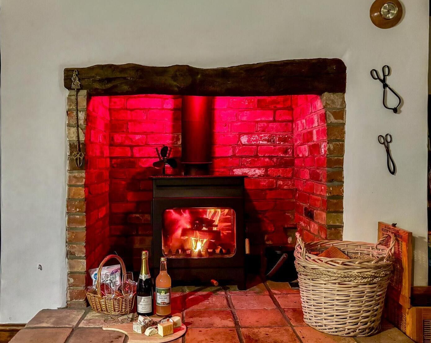 A fireplace with a red brick surround, a wooden mantel, and a fire burning inside. There are bottles and a basket of bread on the floor beside the fireplace.