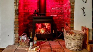 Una chimenea con un marco de ladrillo rojo, un fuste de madera y fuego encendido en su interior. Hay botellas y una cesta de pan en el suelo, al lado de la chimenea.